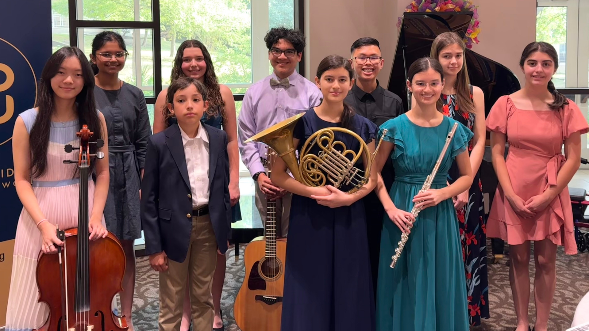 A group of nine young musicians pose together indoors holding their instruments, including a cello, French horn, guitar, and flute, with a grand piano in the background.