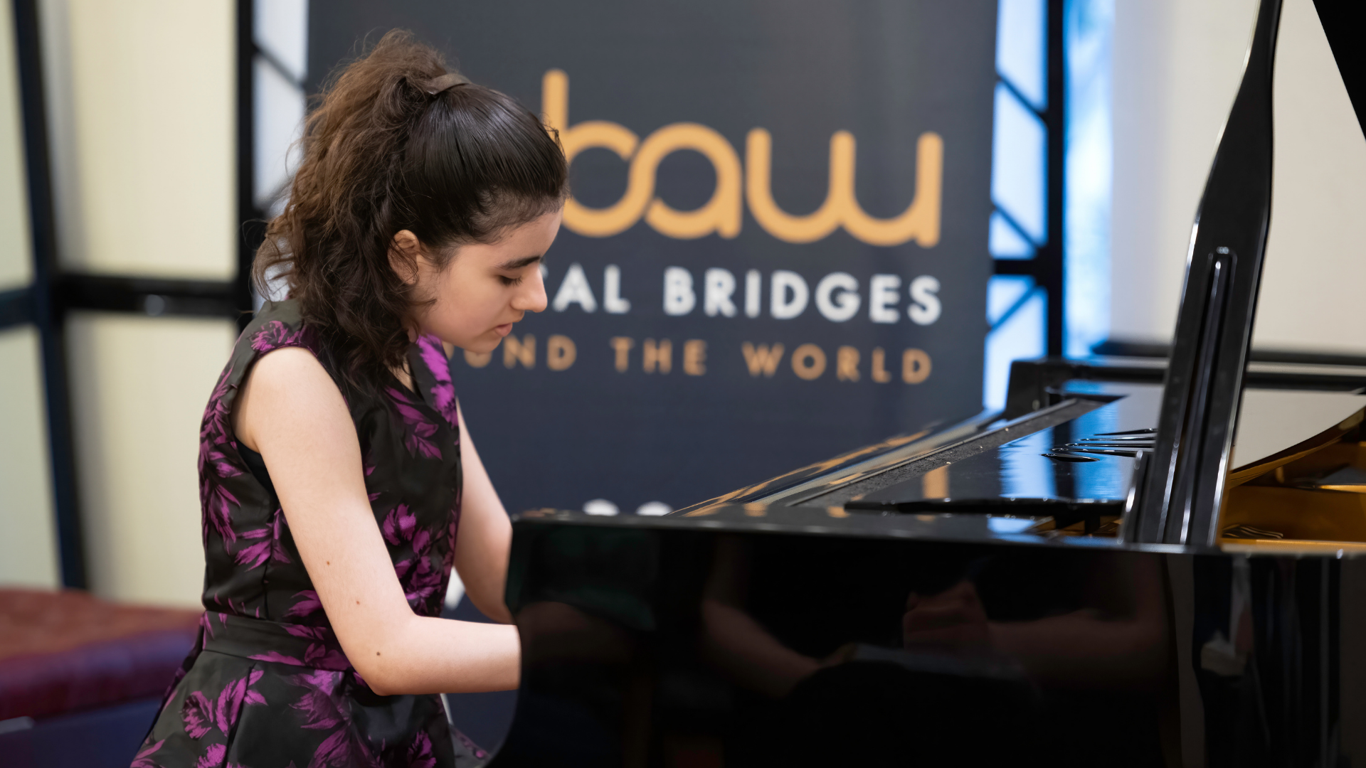 A girl in a black and purple floral dress plays a grand piano, with a Musical Bridges Around the World banner behind her.
