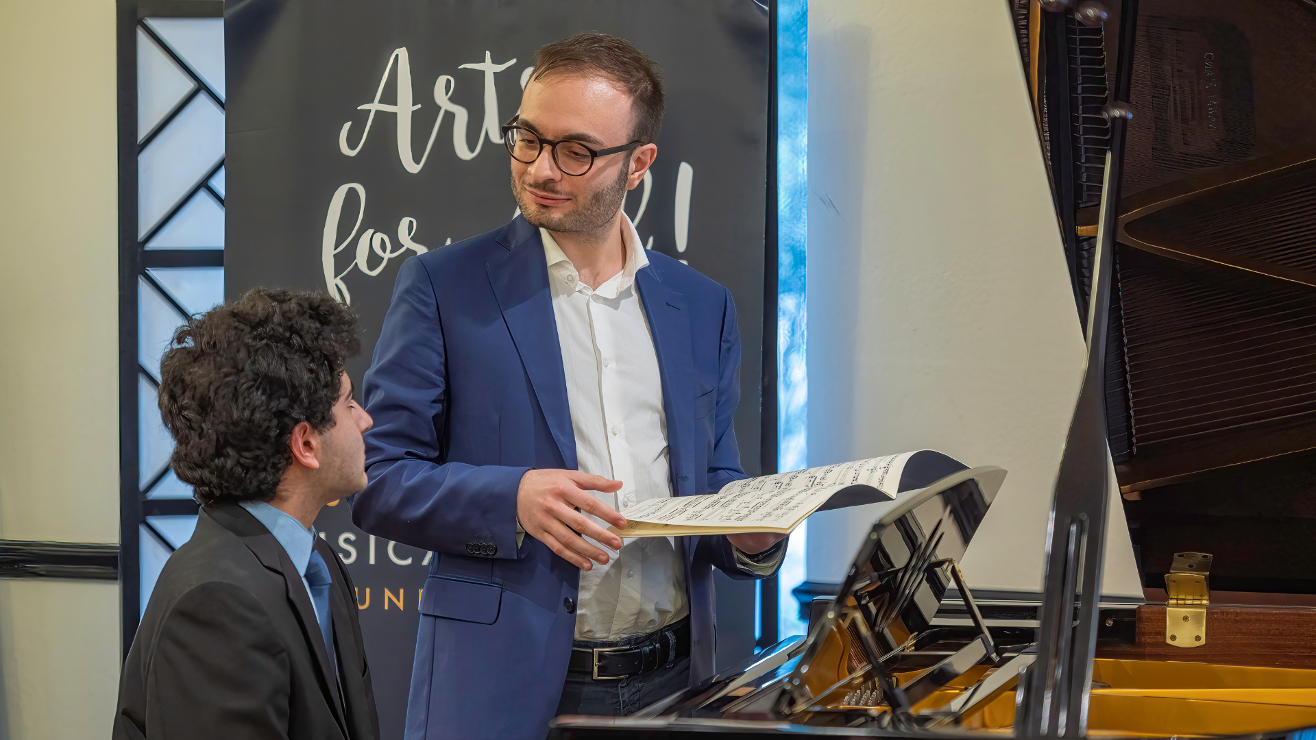 A man in a blue blazer shows sheet music to a seated young man in a suit, standing beside a grand piano with an "Arts for..." banner behind them.
